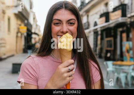 Eine Frau hält einen Eisbecher in der Hand. Sie lächelt und genießt ihre Leckerei. Die Szene spielt auf einer Straße mit mehreren Stühlen Stockfoto