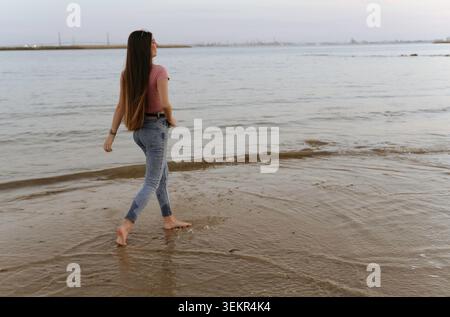 Eine Frau läuft am Strand in einem rosa Hemd und einer blauen Jeans. Das Wasser ist ruhig und der Himmel ist wunderschön blau Stockfoto