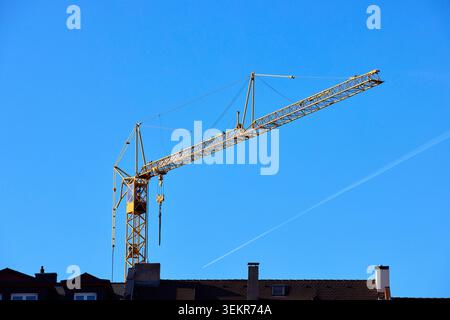 Gelber Turmkran, der auf einer Baustelle über Gebäudedächern vor klarem blauem Himmel eingesetzt wird. Hebegeräte für schwere Industriemaschinen. Stockfoto