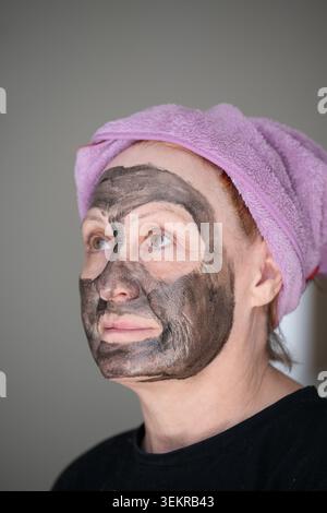 Profilporträt einer Reifen Frau mit schwarzer Gesichtsmaske, die nach oben blickt. Frau mittleren Alters, die eine reinigende Aktivkohle-Spa-Maske auf das Gesicht gegen ein Grau aufträgt Stockfoto