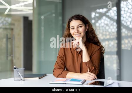 Die lächelnde junge Frau saß an einem Schreibtisch in einem hellen, modernen Büro und legte ihr Kinn auf der Hand mit Notizbüchern und einem Stifthalter im Vordergrund und Glaswänden Stockfoto