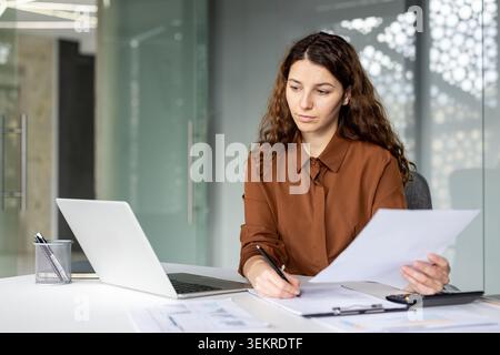 Eine junge Geschäftsfrau sitzt an einem Schreibtisch mit Laptop, Papieren und Zwischenablage und schreibt Notizen, während sie Dokumente in einer modernen Büroumgebung überprüft Stockfoto