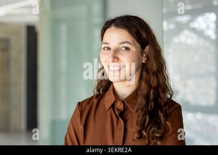 Freundliche junge Frau mit langen welligen dunklen Haaren lächelt in einer hellen, modernen Innenumgebung mit weichem Tageslicht und verschwommenem Hintergrund zur Kamera Stockfoto