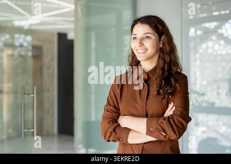 Selbstbewusste junge Frau lächelt, während sie mit gekreuzten Armen in einem hellen, modernen Bürobereich mit Glaswänden und zeitgenössischem Innendesign steht Stockfoto
