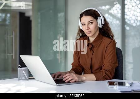 Junge Frau, die mit weißen Kopfhörern an einem Schreibtisch sitzt und auf einem Laptop in einem hellen, modernen Büro mit Glaswänden und einem Tablet in der Nähe schreibt Stockfoto