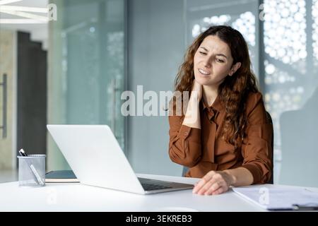Eine junge Frau, die an einem Schreibtisch in einem modernen Büro sitzt und ihren Hals mit angespanntem Ausdruck hält, während sie neben einem offenen Laptop und Papierkram arbeitet Stockfoto