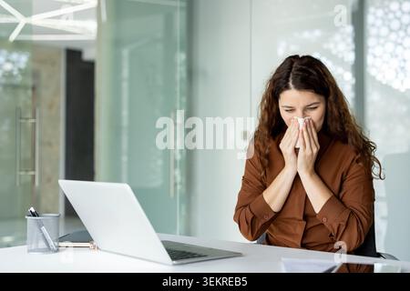 Junge Frau, die an einem Schreibtisch in einem hellen, modernen Büro sitzt und Taschentücher an der Nase hält, während sie neben einem offenen Laptop und Dokumenten arbeitet Stockfoto
