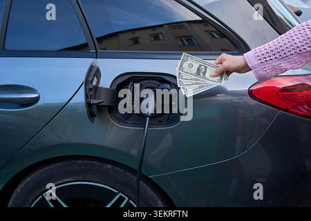 Nahaufnahme einer weiblichen Hand, die US-Dollar-Banknoten neben einem Elektrofahrzeug hält, das an eine Ladestation angeschlossen ist, was die Energiekosten symbolisiert. Stockfoto
