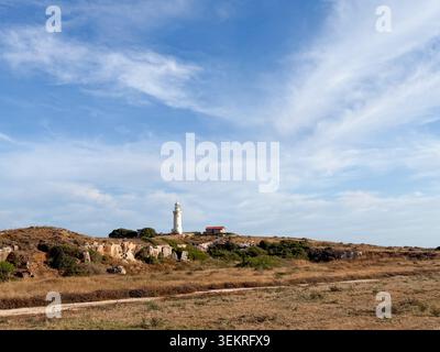 Weitläufige zyprische Landschaft mit gelben sanften Hügeln, die mit trockenem Gras bedeckt sind, einem alten weißen Leuchtturm in der Ferne und einem hellen klaren blauen Himmel Stockfoto