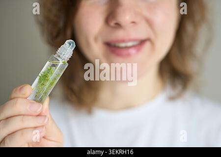 Frau Hält Glasrolle Flasche Mit Grünen Kräutern Nahe Gesicht Lächelnd In Neutral Indoor Closeup Trägt Weißes Hemd Zeigt Hand Und Fingernägel In Weiches Natürliches Licht Häusliche Einstellung. Stockfoto