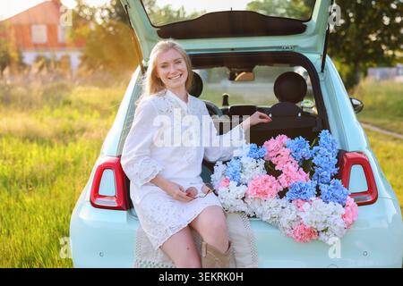 Schöne Frau in einem weißen Kleid posiert mit bunten Hortensien in einem offenen Kofferraum und schafft eine traumhafte Sommer-Picknick- und Urlaubsszene Stockfoto