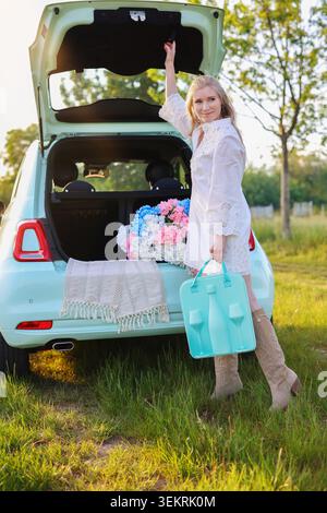 Lächelnde blonde Frau in einem weißen Kleid und Cowboystiefeln, die neben einem offenen, pastellfarbenen Auto steht, das bei Sonnenuntergang auf einer ländlichen Wiese mit bunten Hortensien dekoriert ist Stockfoto