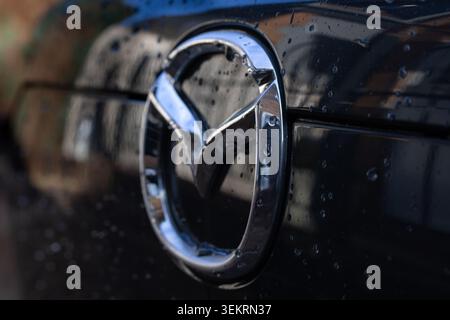 Nahaufnahme des Mazda-Fahrzeugabzeichens mit Regentropfen auf der glänzend schwarzen Karosserie und geringer Schärfentiefe. Stockfoto