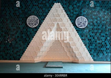 Verzierter Mosche-Mihrab mit wand aus blaugrünen sechseckigen Fliesen, geschnitzter weißer geometrischer Nische und runden Plaketten mit arabischer Kalligraphie in der Moschee. Stockfoto