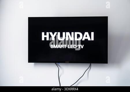 Ein Hyundai Smart LED-Fernseher ist an einer weißen Wand montiert und zeigt seinen leuchtend weißen Startbildschirm mit schwarzen Kabeln darunter. Stockfoto