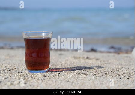 Traditioneller türkischer Tee in einem Tulpenglas an einem Sandstrand mit verschwommenem Meer im Hintergrund, natürliches Sonnenlicht Stockfoto