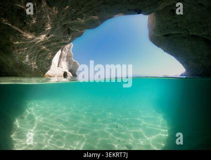Blick auf zwei Ebenen in eine Meereshöhle: Türkisfarbenes klares Wasser, sonnendurchfluteter Sandboden und Felsbogen, der sich zum blauen Himmel öffnet, ideal für Reisen, Sommer und Strand Stockfoto