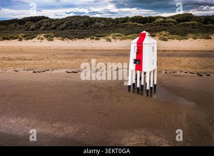 Drohnenblick auf den Burnham‑on‑Sea Low Lighthouse am Sandstrand, der seine markante rot-weiße Struktur vor Dünen und Himmel einfängt Stockfoto