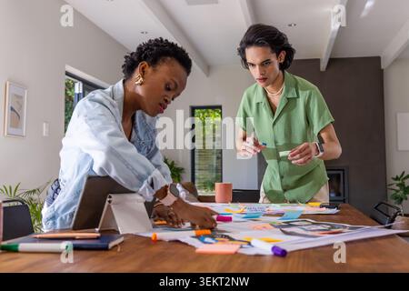 Verschiedene Kollegen, die zu Hause am Tisch stehen und Layouts, Tablets und Haftnotizen arrangieren Stockfoto