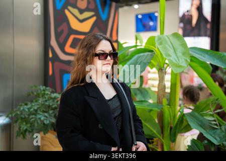 Junge kaukasische Frau mit Sonnenbrille steht in der Nähe tropischer Pflanzen in einem modernen Einkaufszentrum mit Geschäften und dekorativen Elementen. Lebensstil im Einzelhandel Stockfoto