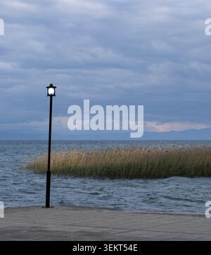 moody seeblick mit einer alten Straßenlaterne auf einem Betonpier unter bewölktem Himmel, mit goldenem Schilf, das sich im welligen Wasser und weit entfernten m bewegt Stockfoto
