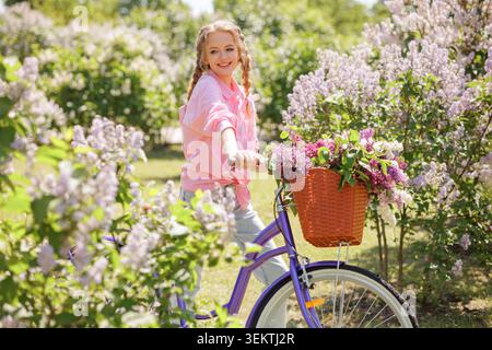 Eine fröhliche junge Frau fährt mit dem Fahrrad durch einen blühenden Blumengarten und zeigt die Freude des Radfahrens und der aktiven Erholung an einem sonnigen Tag Stockfoto