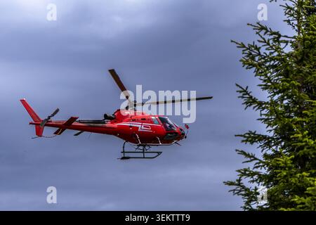 Ein leuchtend roter Hubschrauber schwingt durch einen bewölkten Himmel über einer üppigen, grünen Berglandschaft. Der Helikopter wird während des Fluges gefangen genommen und zeigt seine Powe Stockfoto