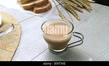 Auf einer rustikalen weißen Holzplatte steht ein Glasbecher mit Schaumkaffee, gefüllt mit handwerklich hergestellten Brotscheiben und dekorativen getrockneten Weizen Stockfoto