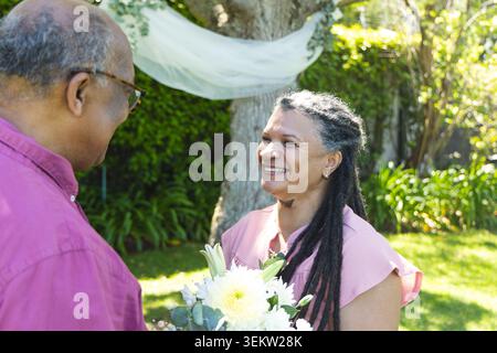 Afroamerikanisches Paar lächelnde Frau mit Blumenstrauß in rosafarbenem Oberteil in der Nähe von Vorhängen im Garten Stockfoto