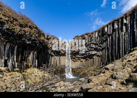 Wasserfall Svartifoss, der über Basaltsäulen mit kleinem Regenbogen unter blauem Himmel in Skaftafell South Coast Island kaskadiert Stockfoto