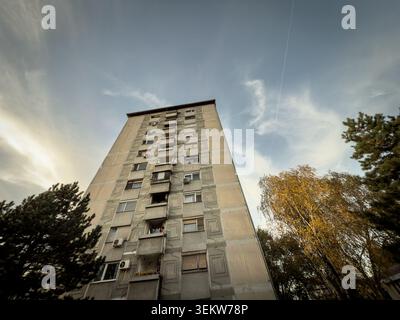 Flacher Blick auf einen brutalistischen Wohnblock in Serbien mit einer Betonfassade aus der kommunistischen Ära, Balkonen, Klimaanlagen und Bäumen Stockfoto