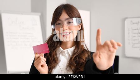 Lächelnde Geschäftsfrau, die eine intelligente Brille trägt und eine Kreditkarte hält, während sie im modernen Büro den virtuellen Bildschirm berührt, Unternehmerin, die Augmente verwendet Stockfoto