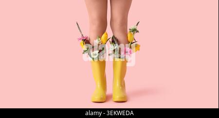 Frauenbeine in gelben Regenstiefeln gefüllt mit bunten Frühlingsblumen auf rosa Hintergrund, kreatives Mode Stillleben mit Tulpen und Gerbera Gänseblümchen, q Stockfoto