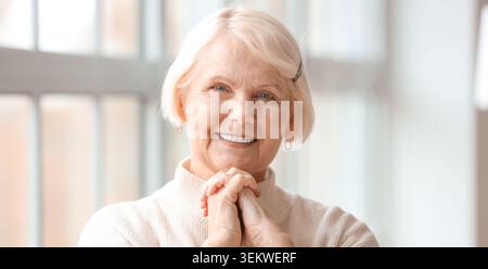 Lächelnde Seniorenporträt drinnen am Fenster, glückliche ältere Frau mit kurzen grauen Haaren und gefalteten Händen, selbstbewusste Rentnerin, die ansah Stockfoto