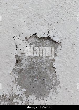 Eine Nahaufnahme der abblätternden, abgebrochenen weißen Farbe, die sich von einer Betonaußenwand ablöst, wodurch der rohe, graue, strukturierte Putz darunter durch Feuchtigkeit freigelegt wird Stockfoto