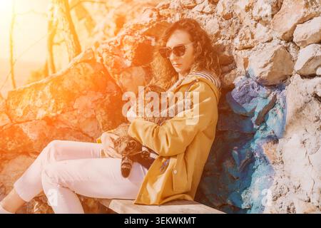 Frau Katze draußen, die auf einer Holzbank sitzt und ein Haustier in warmem goldenem Sonnenlicht an eine Steinmauer hält Stockfoto