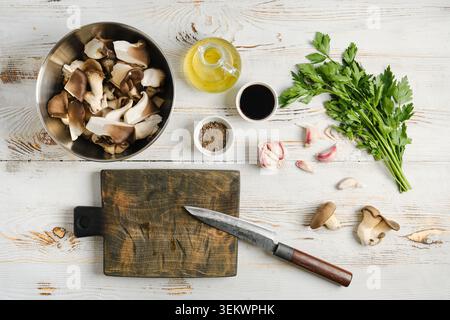 Austernpilze in einer Schüssel zusammen mit Kochöl, Knoblauchzehen und frischen Kräutern. Schneidebrett mit Messer steht bereit für die Zubereitung in einer Küchenumgebung Stockfoto