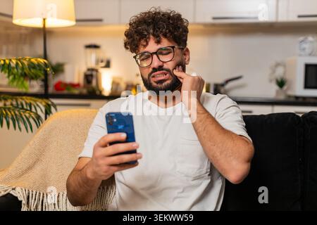 Ein arabischer Mann zu Hause hält Wange mit Zahnschmerzen sucht Zahnarzt auf Smartphone findet Klinikzeit. Ägyptischer Mann auf Sofa fühlt, dass Schmerz runzelt Arten Nachrichten Anfragen Besuchen Sets Termin sucht Schmerzlinderung. Stockfoto