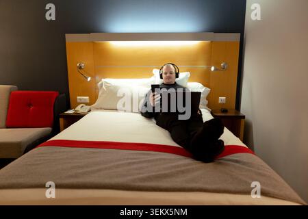 Erwachsener Mann, der in einem Hotelzimmer arbeitet oder sich entspannt und Inhalte auf Telefon und Laptop ansieht Stockfoto