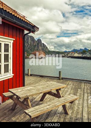 Svolvær, Norwegen - 08.07.2020: Rustikaler Holztisch auf einer Uferterrasse neben einer roten Fischerhütte mit Bergen unter bewölktem Himmel Stockfoto