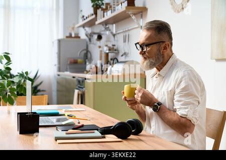 Ein älterer Mann mit Bart sitzt an einem Tisch, schlürft Kaffee und genießt die Ruhe zu Hause. Stockfoto