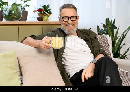 Ein älterer Mann mit Bart entspannt sich in seiner stilvollen Wohnung, während er ein warmes Getränk schlürft. Stockfoto
