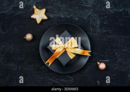 Schwarzer Teller mit Geschenkbox mit goldener Schleife und Weihnachten auf dunklem strukturiertem Hintergrund. Urlaubskonzept. Draufsicht, flach. Stockfoto