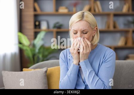 Reife Frau, die zu Hause auf einem Sofa sitzt, ihre Nase in ein Gewebe bläst, an saisonalen Allergien, einer Erkältung oder Grippe leidet und Beschwerden und Krankheiten erlebt Stockfoto