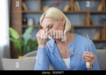 Reife Frau mit kurzen blonden Haaren, die Schläfen reiben und Brillen halten, zu Hause an Migräne und Augenanstrengung leidet, müde, gestresst und mit sichtbaren Schmerzen und Beschwerden Stockfoto