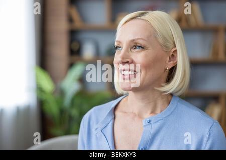 Reife Frau mit blonden Haaren und blauen Augen, die warm lächelt und Glück und Optimismus in einer komfortablen Innenumgebung ausdrückt, die Wohlbefinden und positive Emotionen repräsentiert Stockfoto