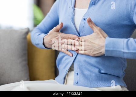 Frau, die auf einem Sofa sitzt und unter starken Schmerzen in der Brust oder Sodbrennen leidet, ihre Hände auf die Brust legt, was auf Unbehagen, gesundheitliche Probleme und medizinische Symptome hinweist Stockfoto