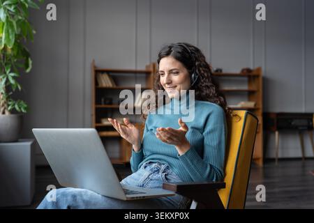 Positiv lächelnde weibliche Mitarbeiterin im Home Office, die ein Headset trägt, Videokonferenz mit dem Kunden, zufriedene Geschäftsfrau, die auf den Laptop schaut und online mit dem Team über das Internet kommuniziert Stockfoto