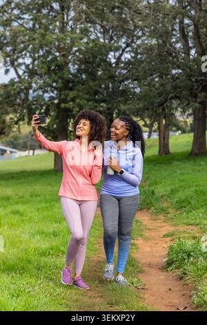 Verschiedene Freundinnen laufen in Sportbekleidung auf dem Parkweg und machen Selfie mit Telefon in der Flasche. Begleiter, Outdoor, Athleisure, Greenspace, Trail, Fitness, Freundschaft Stockfoto