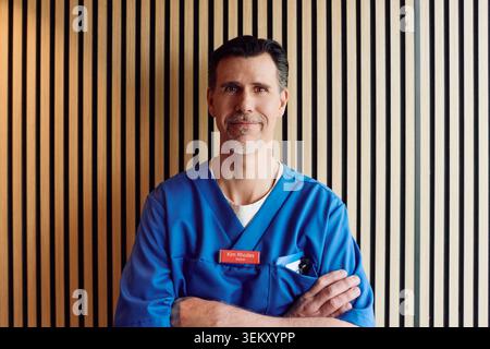 Porträt eines lächelnden männlichen Facharztes mit gekreuzten Armen an der gestreiften Wand im Krankenhaus Stockfoto